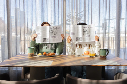 Two female presenting people reading newspapers in Colorado crew neck sweatshirts with coffee mugs and breakfast plates