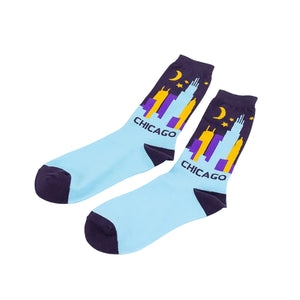 A pair of light blue crew socks with dark purple cuffs, heels, and toes, featuring a colorful Chicago skyline graphic with the word 'CHICAGO'. The graphic includes yellow and purple skyscrapers, a yellow crescent moon, and small yellow stars.