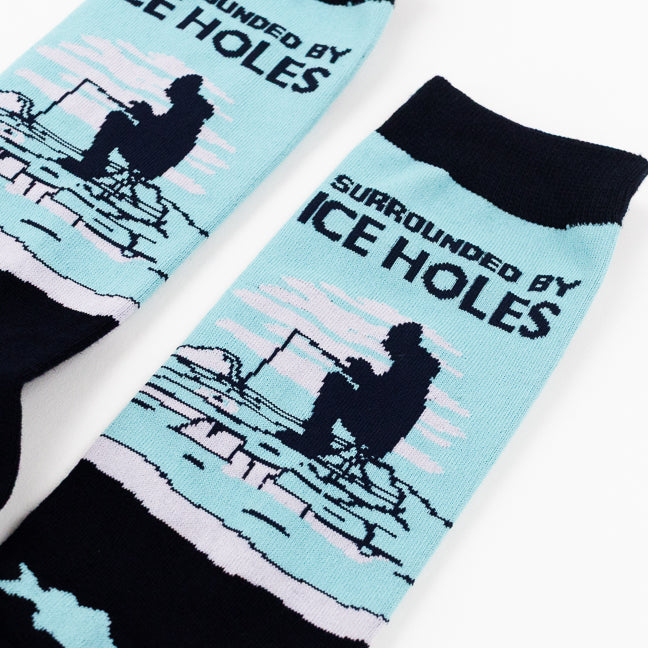 A pair of crew socks on a plain white background. The socks are dark navy with a light blue graphic on the upper calf showing a figure ice fishing with the text 'SURROUNDED BY ICE HOLES', and a lower section with light blue fish and bubbles.