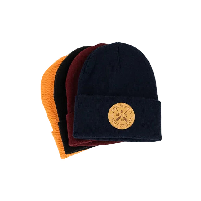 Four knit cuffed beanies stacked slightly offset on a white background, with the front hat in dark navy and a circular tan logo patch on the cuff. The visible hats behind it are orange, black, and burgundy.