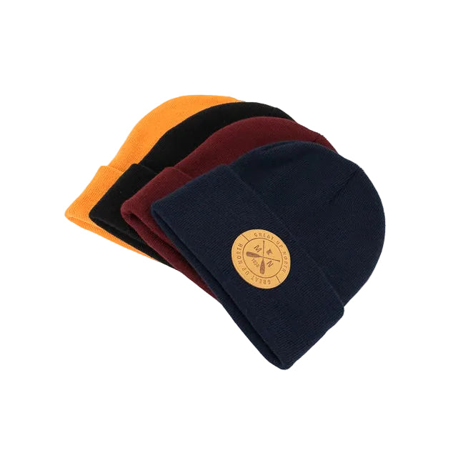 Four knit beanie hats stacked in a fan arrangement on a white background. The hats are navy blue, burgundy, black, and orange, each featuring a circular tan logo patch on the front cuff.