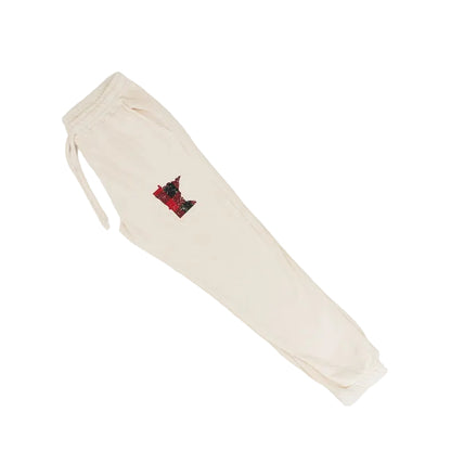 A pair of cream-colored sweatpants with a small red and black embroidered patch on the upper left thigh, laid flat on a white background.