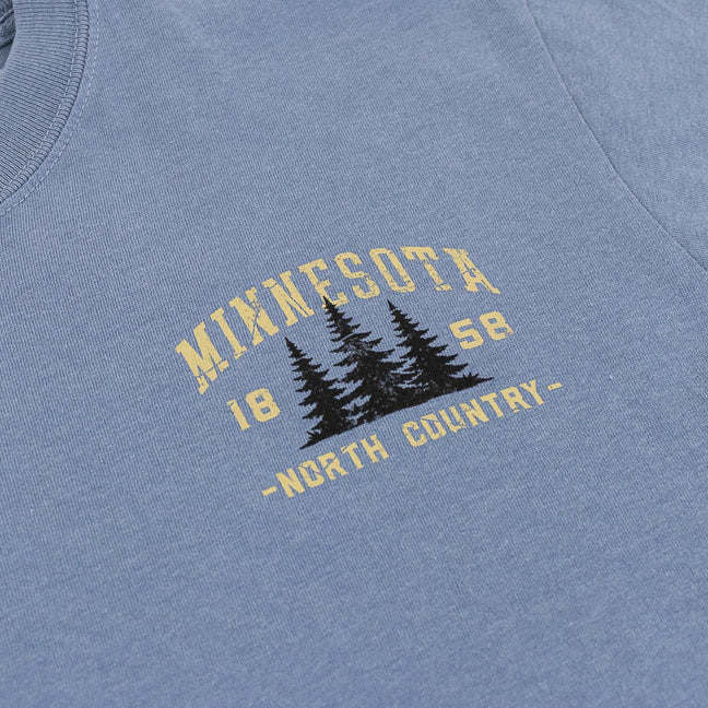 Close-up of a heathered blue t-shirt featuring a vintage-style graphic on the chest with the text 'MINNESOTA 1858 -NORTH COUNTRY-' and three black pine trees.