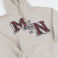 A close-up of a beige pullover hoodie featuring a large collegiate-style 'MN,' the numbers '18' and '58' and a circular seal printed on the chest. The letters 'M' and 'N' are filled with a red and black buffalo plaid pattern.