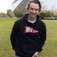 A man wearing a black pullover hoodie with a red and white 'MINNESOTA' graphic, standing outdoors on a grassy lawn with a modern stadium building in the background.