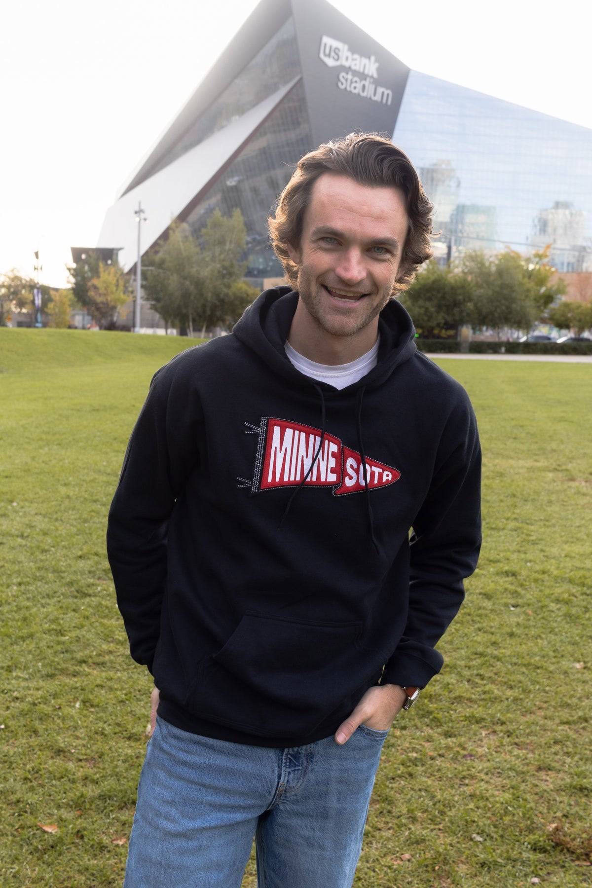 A man wearing a black pullover hoodie with a red and white 'MINNESOTA' graphic, standing outdoors on a grassy lawn with a modern stadium building in the background.