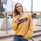 A woman leans against a railing and smiles in front of a football stadium. She wears a yellow sweatshirt with purple text that reads Minnesota. 