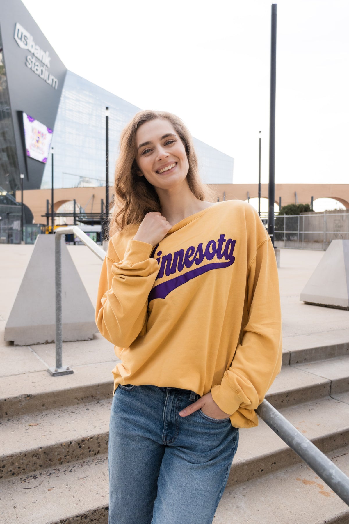 A woman leans against a railing and smiles in front of a football stadium. She wears a yellow sweatshirt with purple text that reads Minnesota. 