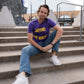 A photo of man sitting on steps in front of a football stadium, wearing a purple shirt with a viking profile and yellow text reading "Defend the North."