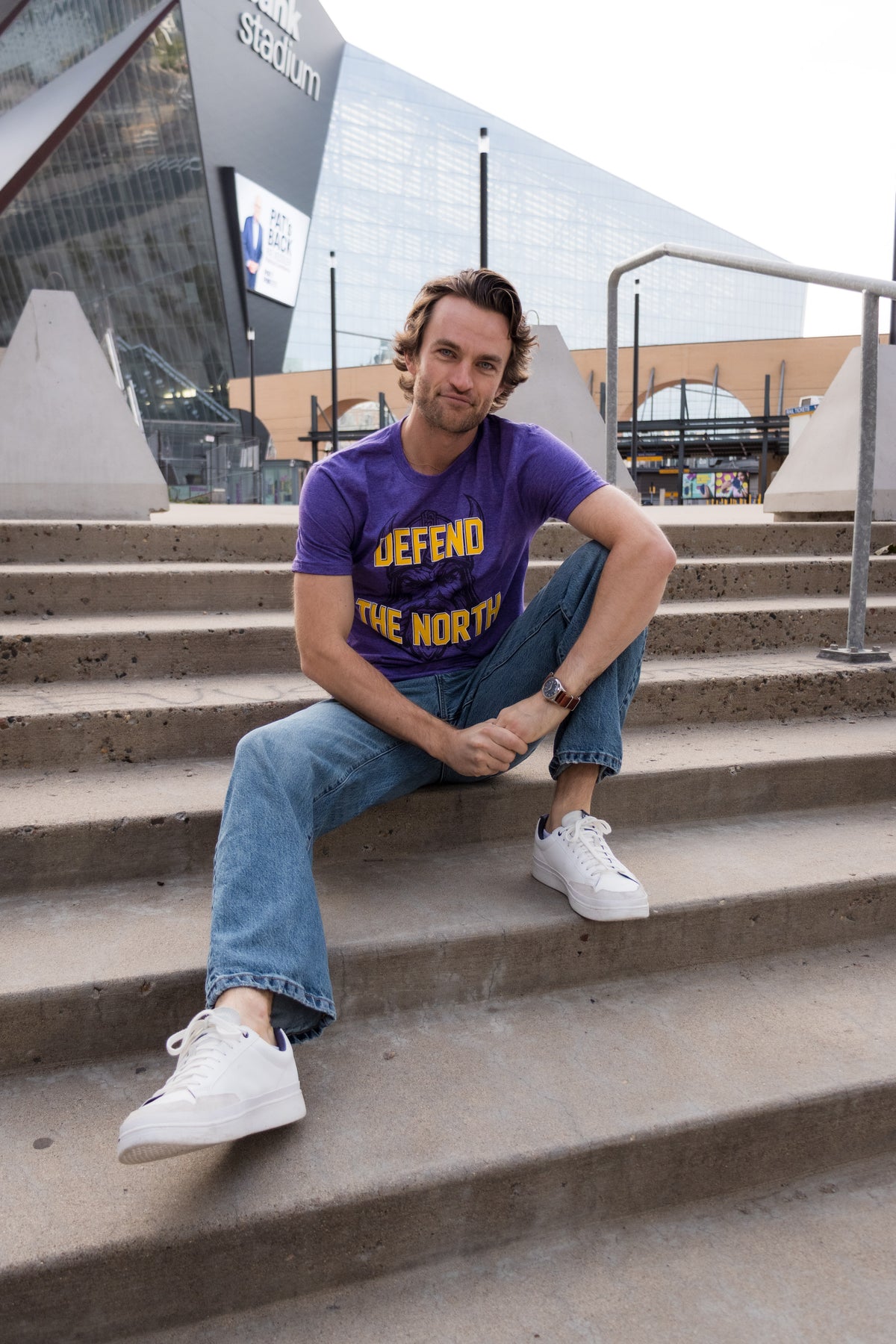 A photo of man sitting on steps in front of a football stadium, wearing a purple shirt with a viking profile and yellow text reading "Defend the North."