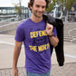 A photo of man sitting on steps in front of a football stadium, wearing a purple shirt with a viking profile and yellow text reading "Defend the North."