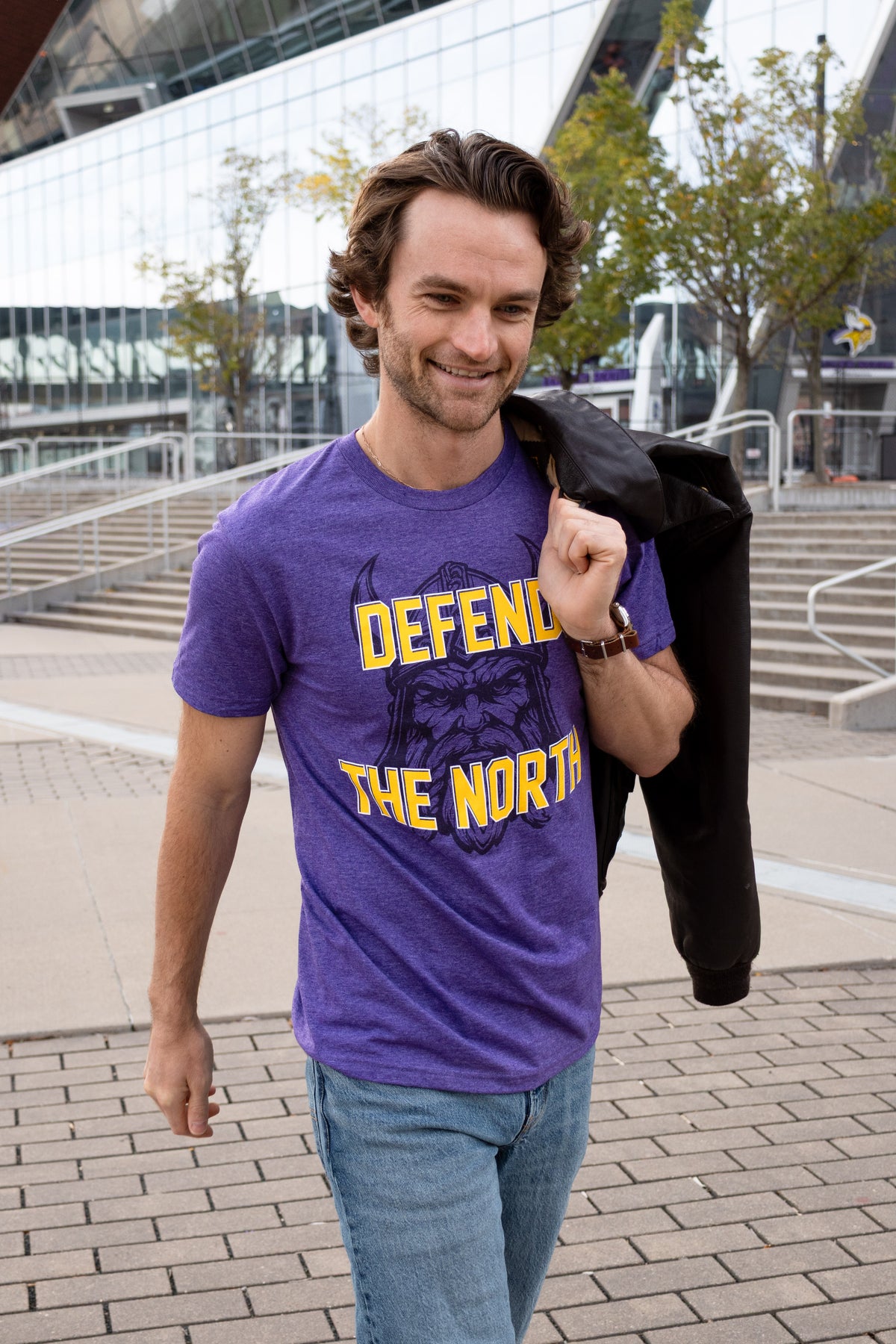 A photo of man sitting on steps in front of a football stadium, wearing a purple shirt with a viking profile and yellow text reading "Defend the North."