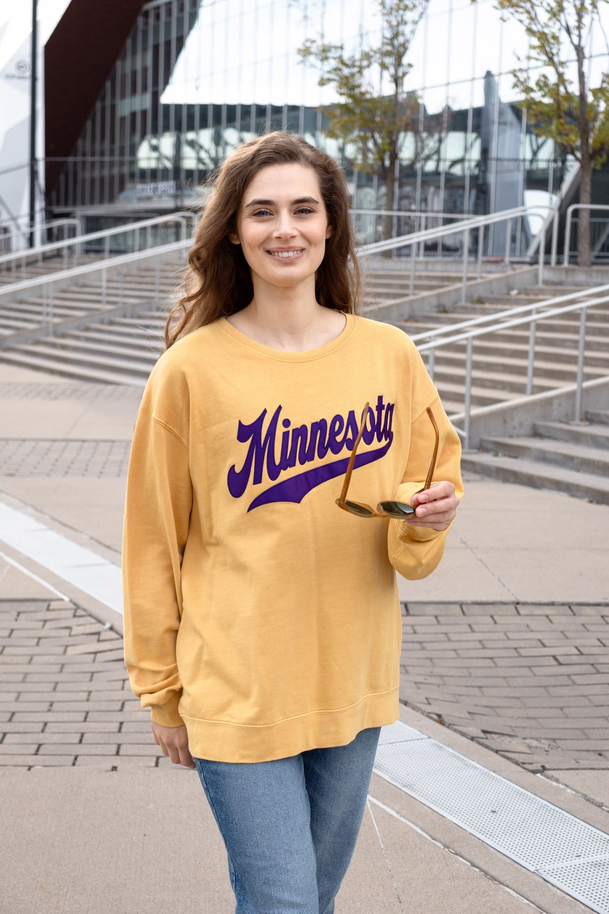 A woman smiles as she walks past a football stadium. She wears a yellow sweatshirt with purple text that reads Minnesota. 
