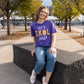A women sits on a large concrete block in a garden outside a large glass building. She wears a purple t-shirt that reads "Let's SKOL Crazy," blue jeans and white tennis shoes. 