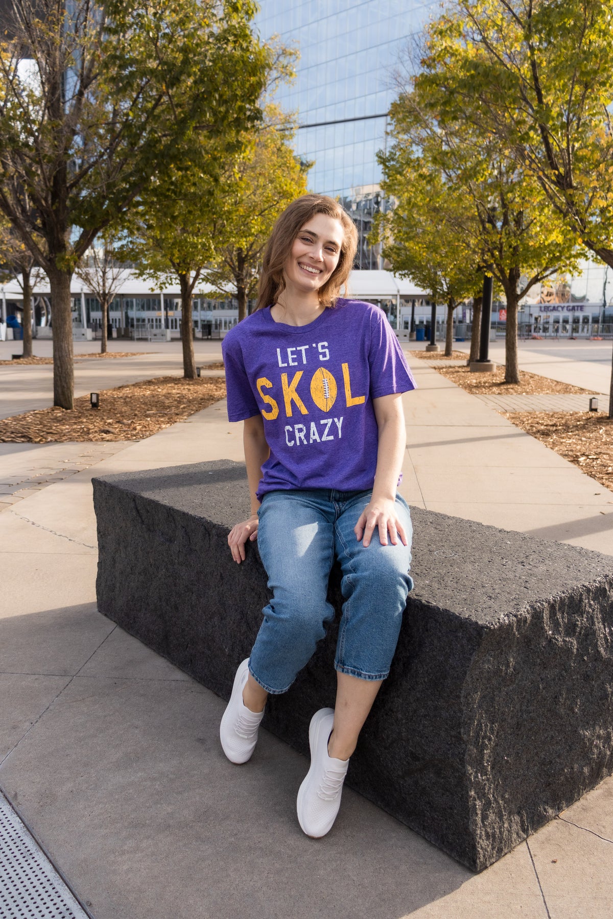 A women sits on a large concrete block in a garden outside a large glass building. She wears a purple t-shirt that reads "Let's SKOL Crazy," blue jeans and white tennis shoes. 