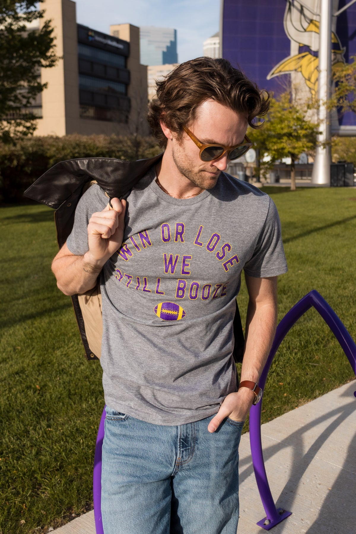 A man wearing a gray graphic t-shirt with purple and yellow text that reads 'WIN OR LOSE WE STILL BOOZE' with a football logo, standing outdoors on a grassy lawn.