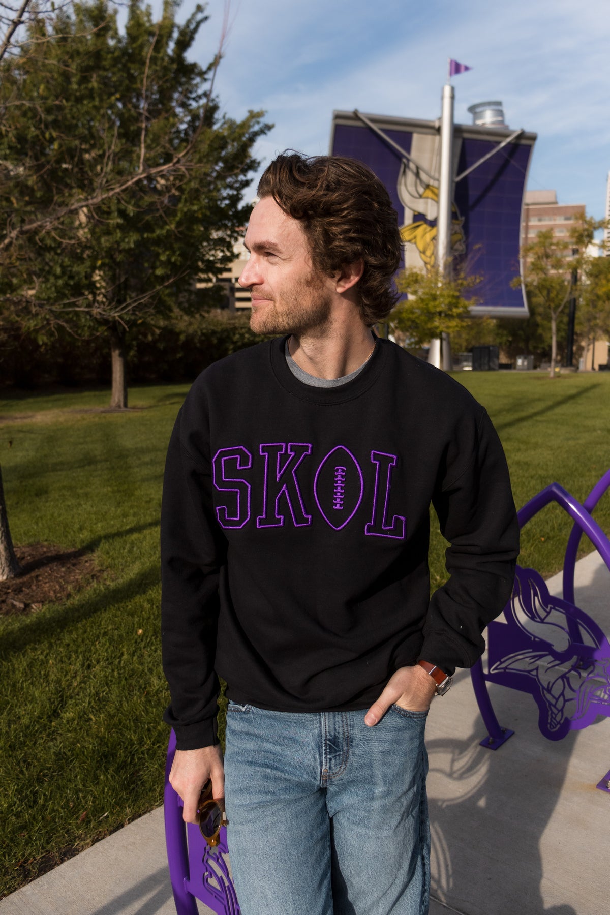 A man stands outside wearing a black sweatshirt with purple letters that reads SKOL. The O in SKOL is in the shape of a football.