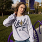 A woman stands outside wearing a light heather grey sweatshirt that reads SKOL, MN, est. 858. A gold compass star replaces the O in SKOL. 