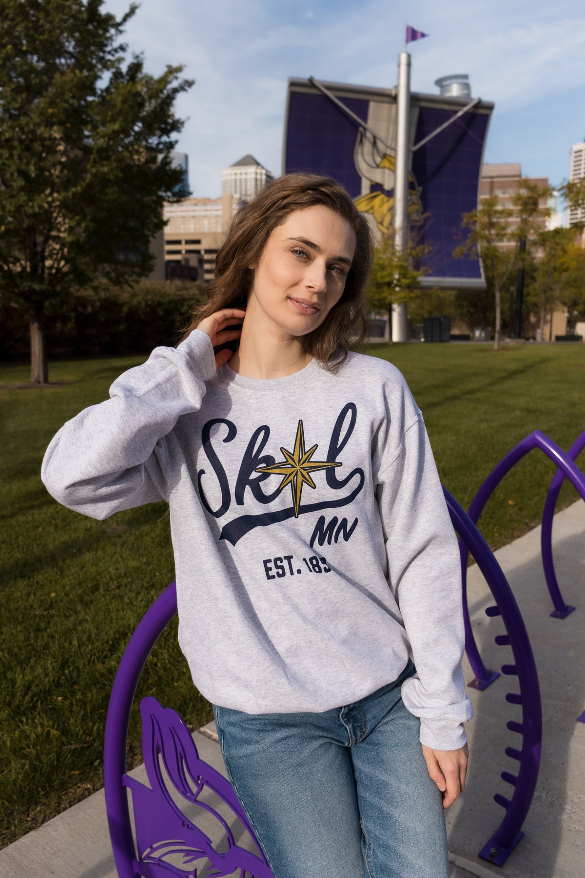 A woman stands outside wearing a light heather grey sweatshirt that reads SKOL, MN, est. 858. A gold compass star replaces the O in SKOL. 