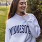 A woman wearing a light heather gray sweatshirt with 'MINNESOTA' printed in navy blue arched letters across the chest, standing outdoors in a park-like setting with green grass and trees.
