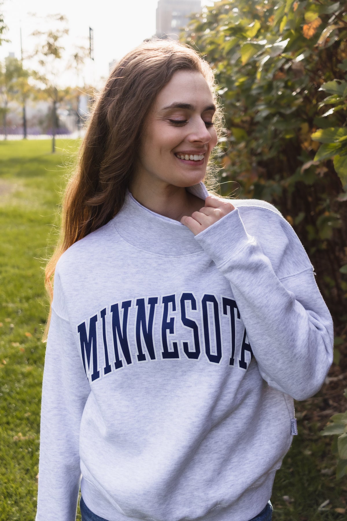 A woman wearing a light heather gray sweatshirt with 'MINNESOTA' printed in navy blue arched letters across the chest, standing outdoors in a park-like setting with green grass and trees.