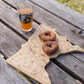 A wooden cutting board shaped like the state of Minnesota, engraved with state outlines and place names, sits on a weathered wooden picnic table outdoors. Two sugar-dusted donuts rest on the board, and a clear drinking glass with an orange beverage and a dark camping-themed graphic is also placed on the table.