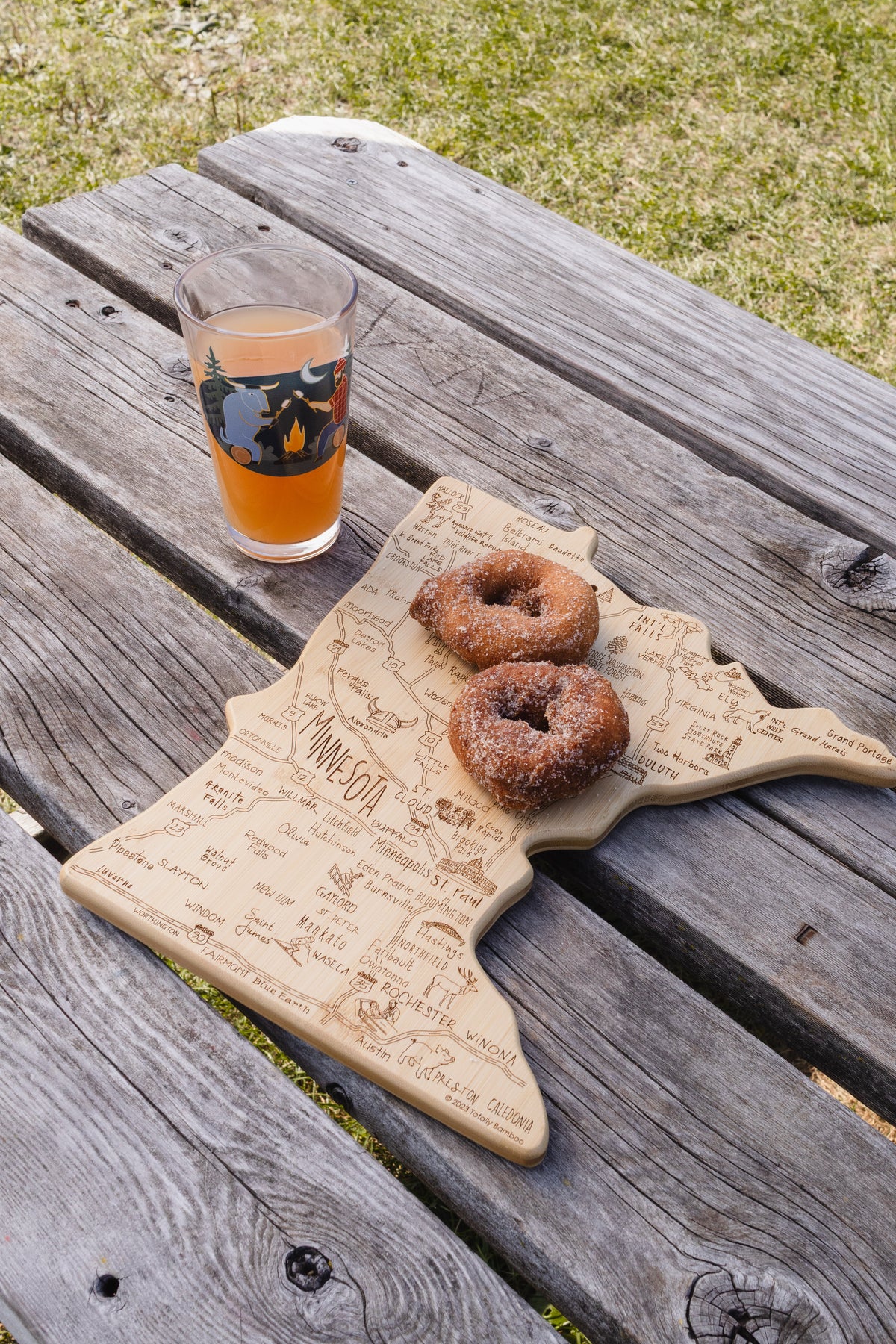 A wooden cutting board shaped like the state of Minnesota, engraved with state outlines and place names, sits on a weathered wooden picnic table outdoors. Two sugar-dusted donuts rest on the board, and a clear drinking glass with an orange beverage and a dark camping-themed graphic is also placed on the table.