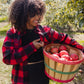 A woman with curly hair stands in an apple orchard holding a wooden basket filled with red apples. She is wearing a red and black plaid flannel shirt over a black top and black leggings.