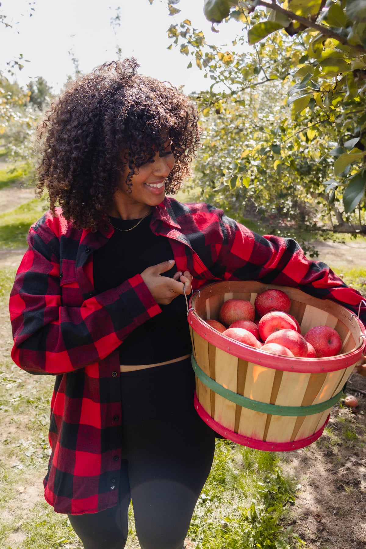 A woman with curly hair stands in an apple orchard holding a wooden basket filled with red apples. She is wearing a red and black plaid flannel shirt over a black top and black leggings.