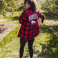 A woman standing in an apple orchard wearing a red and black buffalo plaid flannel shirt with 'MINNESOTA' and a moose graphic printed on the back, paired with black leggings and tan boots.