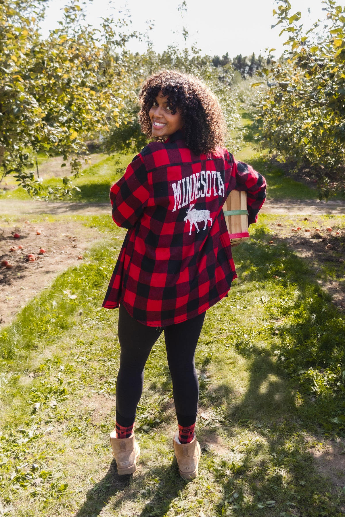 A woman standing in an apple orchard wearing a red and black buffalo plaid flannel shirt with 'MINNESOTA' and a moose graphic printed on the back, paired with black leggings and tan boots.