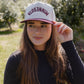 A woman wearing a white and burgundy Minnesota snapback cap, standing outdoors in a sunny field with trees in the background.