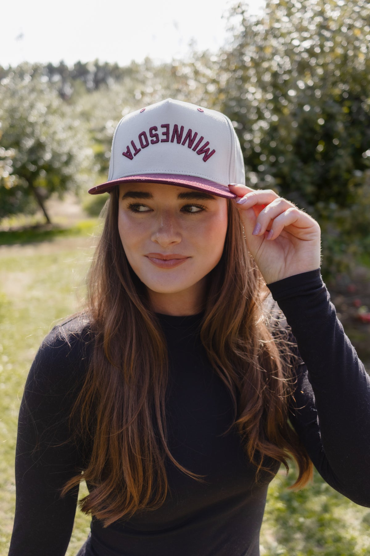 A woman wearing a white and burgundy Minnesota snapback cap, standing outdoors in a sunny field with trees in the background.