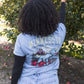 A woman standing outdoors in front of an apple tree, viewed from behind, wearing a light blue graphic T-shirt with 'MINNESOTA LEGENDS NEVER DIE' text and an illustration of Paul Bunyan and Babe the Blue Ox on the back, paired with light blue jeans and black long sleeves.