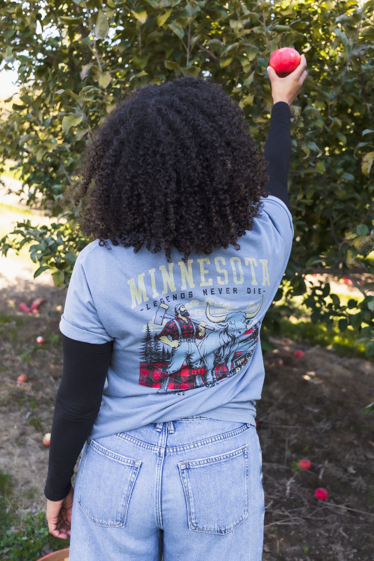 A woman standing outdoors in front of an apple tree, viewed from behind, wearing a light blue graphic T-shirt with 'MINNESOTA LEGENDS NEVER DIE' text and an illustration of Paul Bunyan and Babe the Blue Ox on the back, paired with light blue jeans and black long sleeves.