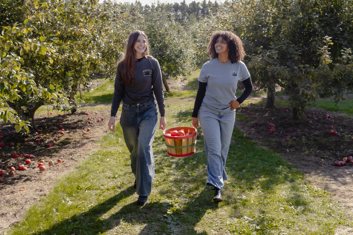 Two women walking through a sunny apple orchard, carrying a basket of red apples. They are wearing matching gray and light blue shirts with a small white logo on the chest, paired with jeans.