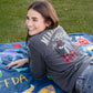 A woman lying on a blue blanket outdoors on a grassy lawn, wearing a dark gray graphic t-shirt and blue jeans.
