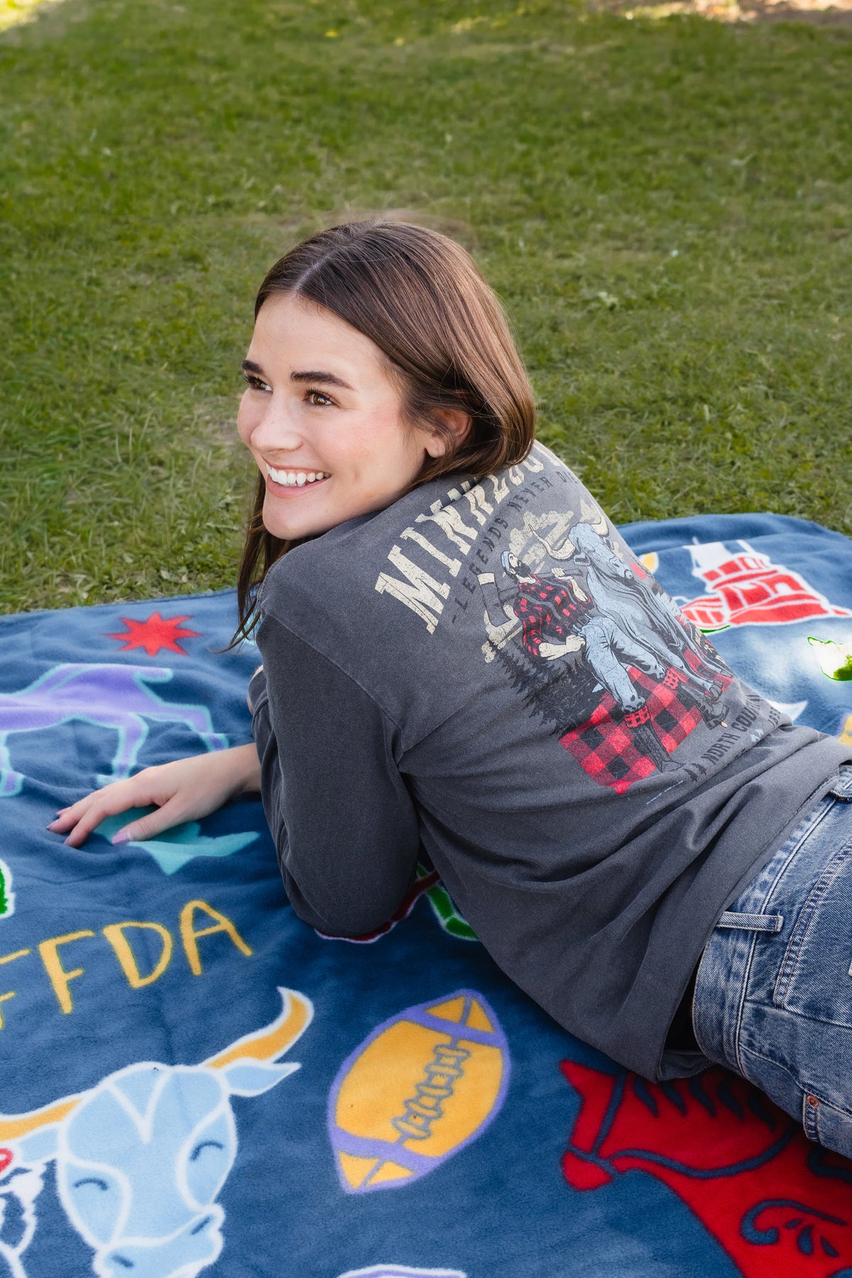 A woman lying on a blue blanket outdoors on a grassy lawn, wearing a dark gray graphic t-shirt and blue jeans.