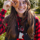 A smiling young woman outdoors in a sunlit orchard, holding a glazed donut over one eye. She wears a red and black buffalo plaid flannel shirt under a black quilted vest.