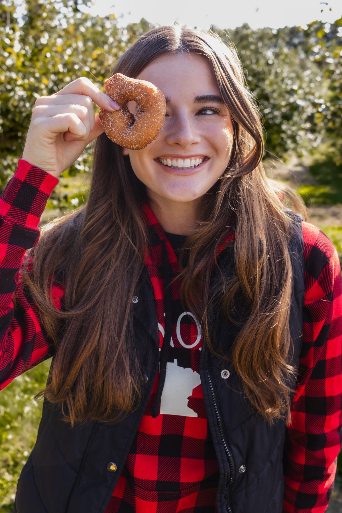 A smiling young woman outdoors in a sunlit orchard, holding a glazed donut over one eye. She wears a red and black buffalo plaid flannel shirt under a black quilted vest.