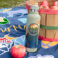 A reusable insulated water bottle with a Minnesota landscape label stands on a blue patterned blanket outdoors, next to a wooden basket of red apples and a single red apple.