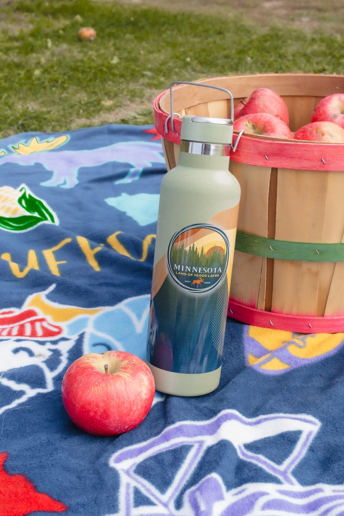 A reusable insulated water bottle with a Minnesota landscape label stands on a blue patterned blanket outdoors, next to a wooden basket of red apples and a single red apple.