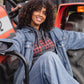 A smiling woman with curly hair sits on an orange tractor, wearing a blue denim jacket over a red and black plaid hoodie and matching blue jeans.
