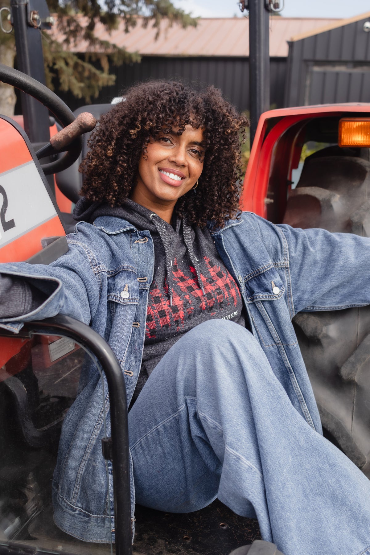 A smiling woman with curly hair sits on an orange tractor, wearing a blue denim jacket over a red and black plaid hoodie and matching blue jeans.