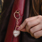 A close-up of a person's hand holding a gold heart-shaped padlock keychain with rhinestones and the word 'MINNESOTA' engraved on it, attached to a red leather shoulder bag.