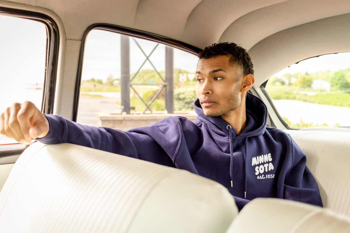 A man sitting in the back of a vintage car wearing a navy blue hoodie with white text that reads 'Minnesota Et. 1858'