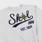 A close-up of a light heather gray sweatshirt featuring a graphic print of the word 'Skol' in dark navy script with a gold compass star over the 'o', 'MN' in dark navy, and 'EST. 1858' below.