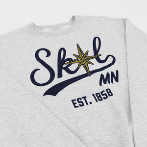 A close-up of a light heather gray sweatshirt featuring a graphic print of the word 'Skol' in dark navy script with a gold compass star over the 'o', 'MN' in dark navy, and 'EST. 1858' below.