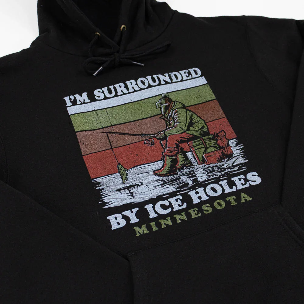 A black sweatshirt featuring a wintry ice fishing scene. Text reads "I'm surrounded by Ice Holes" and "Minnesota."