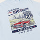 Light blue t-shirt with a graphic of a red car, Route 66 sign, and text celebrating 100 years on a beige background.